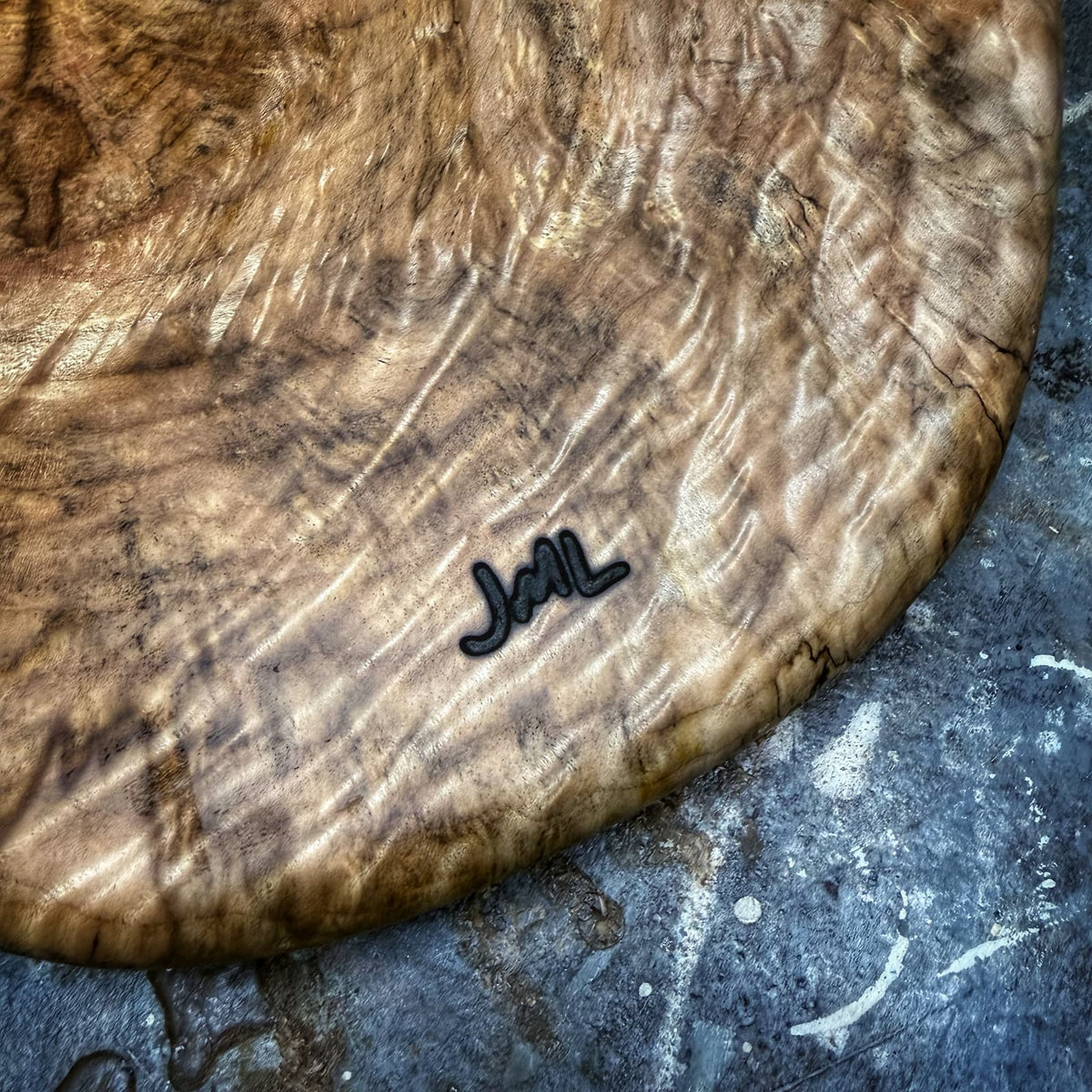 A close-up of a large, round wooden serving board with dramatic wavy grain patterns and a small black metal 'JML' logo, resting on a textured gray stone surface.