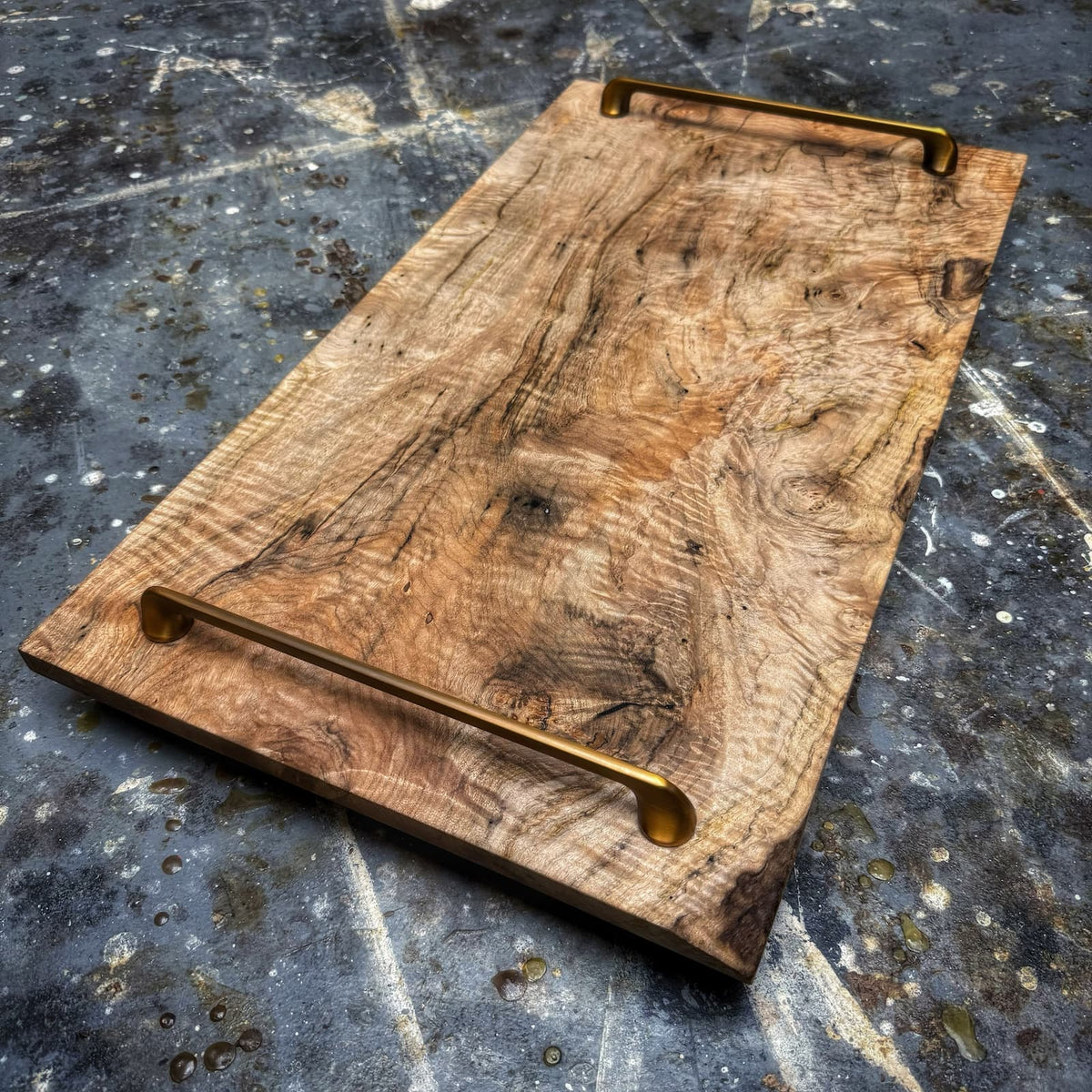 A rustic rectangular wooden serving board with a natural wood grain surface and two brass metal handles, shown on a dark textured stone surface.