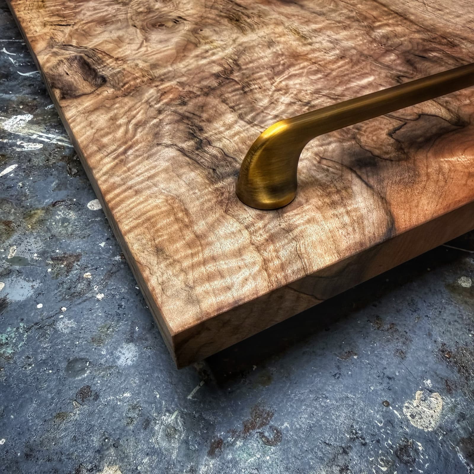 A close-up of a rectangular wooden board or tray with dramatic natural wood grain, featuring a polished brass handle and resting on a textured gray surface.