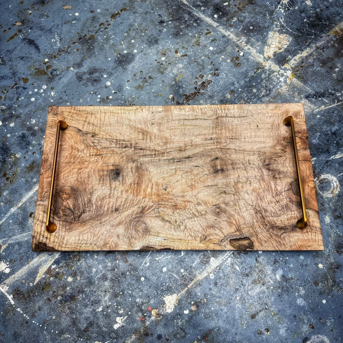 A rustic rectangular wooden serving board with a natural wood grain surface and two brass handles on the left and right sides, placed on a textured gray concrete surface.