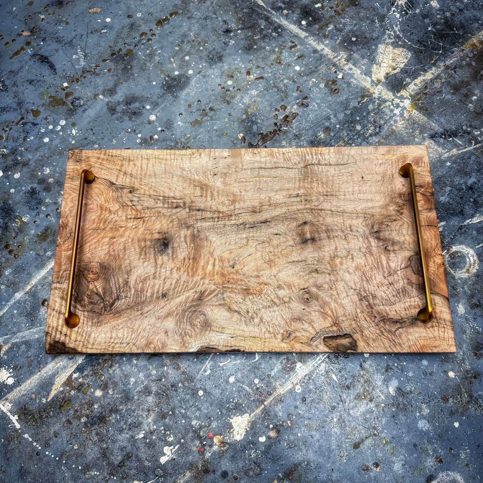 A rustic rectangular wooden serving board with a natural wood grain surface and two brass handles on the left and right sides, placed on a textured gray concrete surface.