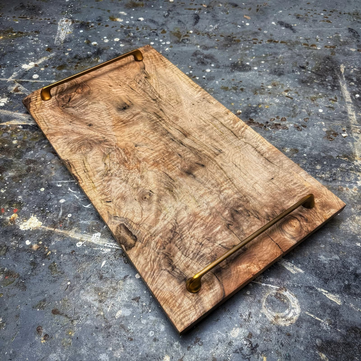 A rustic rectangular wooden serving board with a natural wood grain surface and two brass metal handles, shown on a textured gray concrete floor.