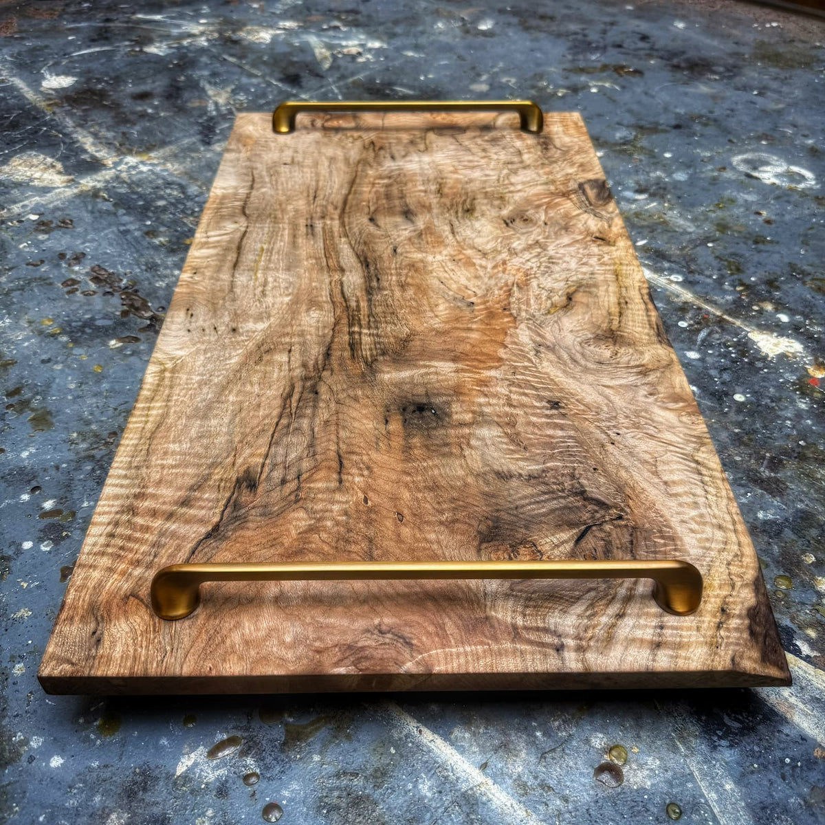 A rectangular rustic wooden serving board with a natural grain pattern and two brass metal handles, one at the top and one at the bottom, resting on a textured gray concrete surface.