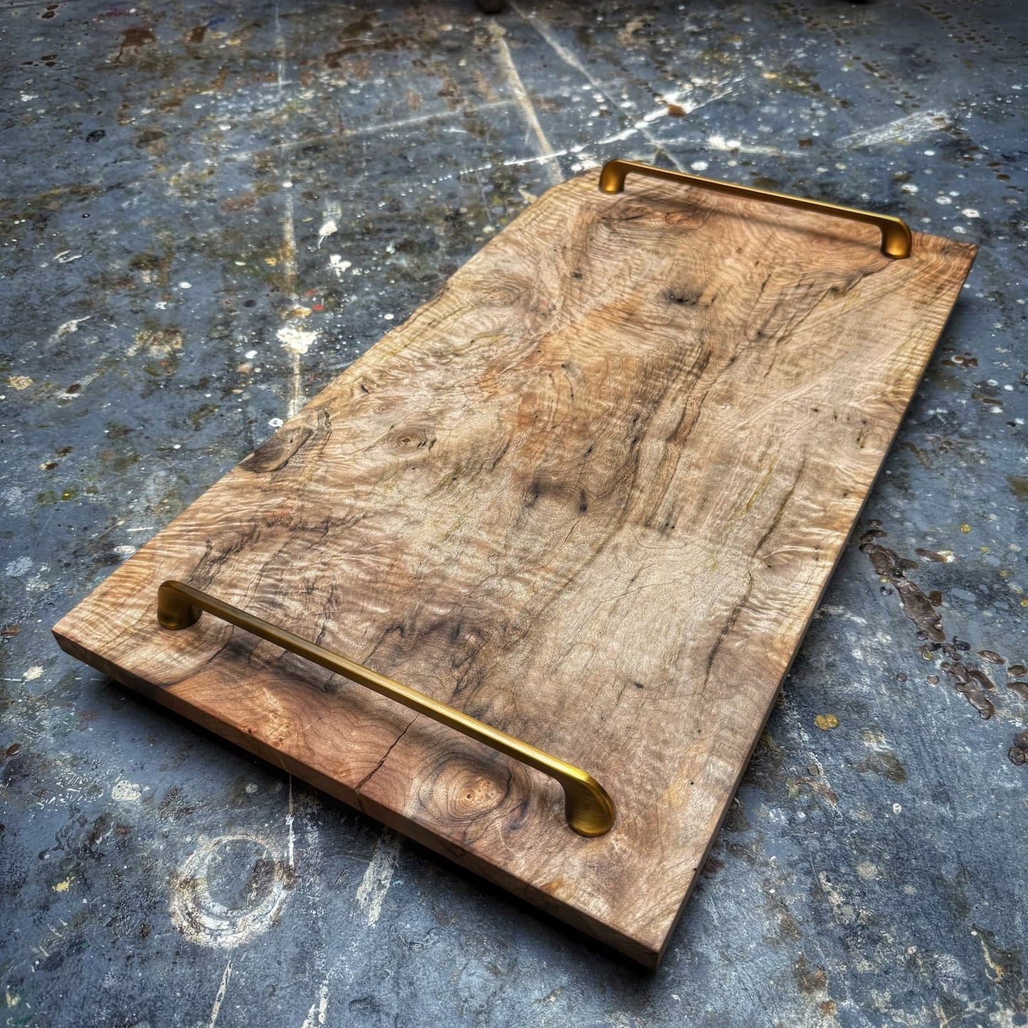 A rustic rectangular wooden serving board with two long brass handles, photographed on a weathered gray concrete surface.