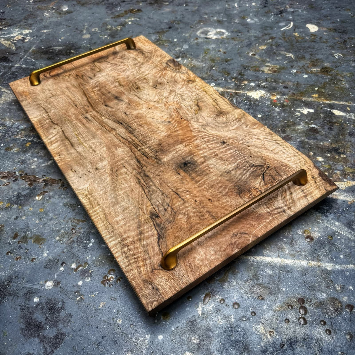 A rustic rectangular wooden serving board with a natural wood grain pattern and two brass handles, shown on a textured concrete floor.