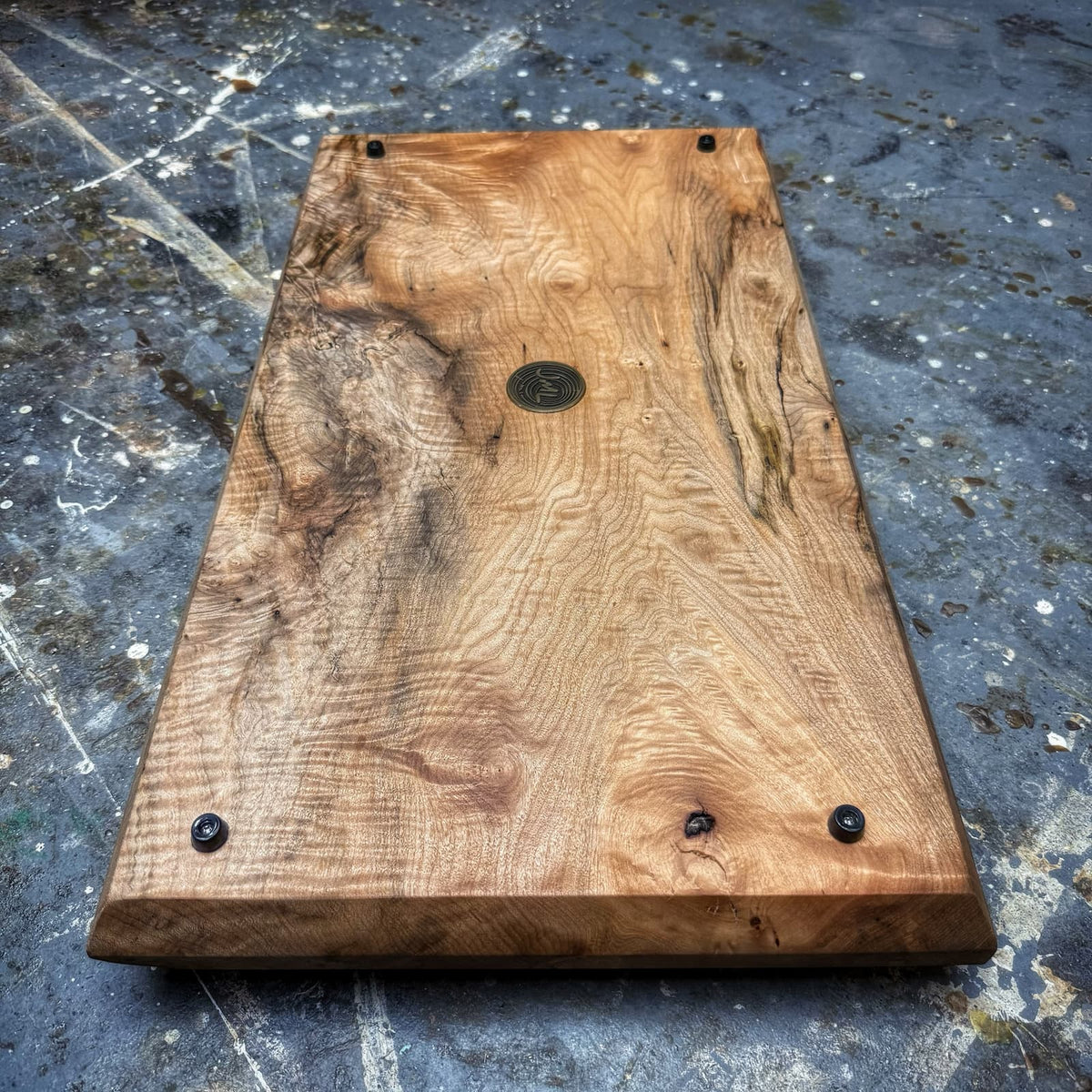 A rectangular handcrafted wooden board with dramatic natural grain patterns, dark metal screws at the corners, and a round metal emblem in the center, photographed on a textured gray concrete floor.