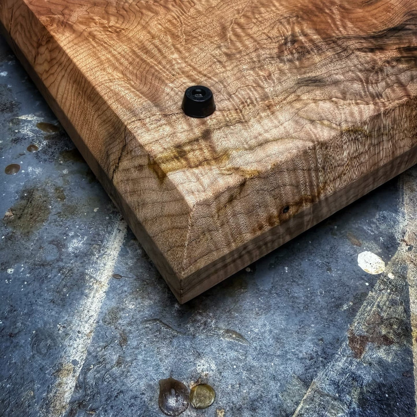 A close-up of a thick, polished wooden cutting board with dramatic curly grain, featuring a black plastic drop weight on top, set on a textured gray concrete floor.