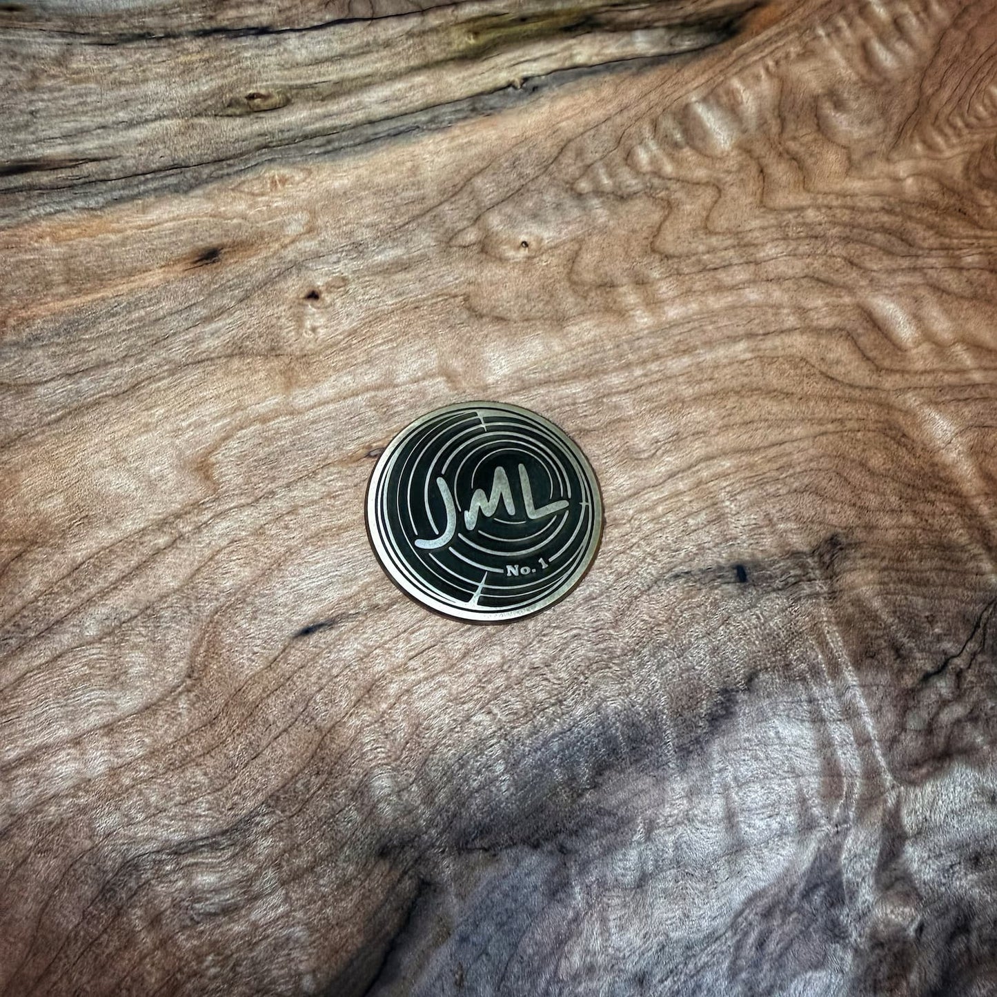 A round black and silver metal coin or medallion with a stylized 'JML' logo and 'Nov. 1' text, placed on a textured, highly figured wood surface.