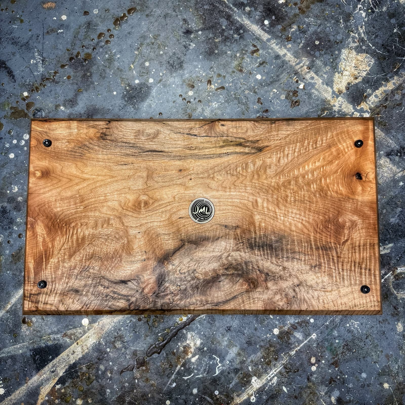 A rectangular handcrafted wooden board with a rich natural grain pattern, secured with black metal grommets at the corners and a circular metal logo in the center, displayed on a textured gray concrete surface.