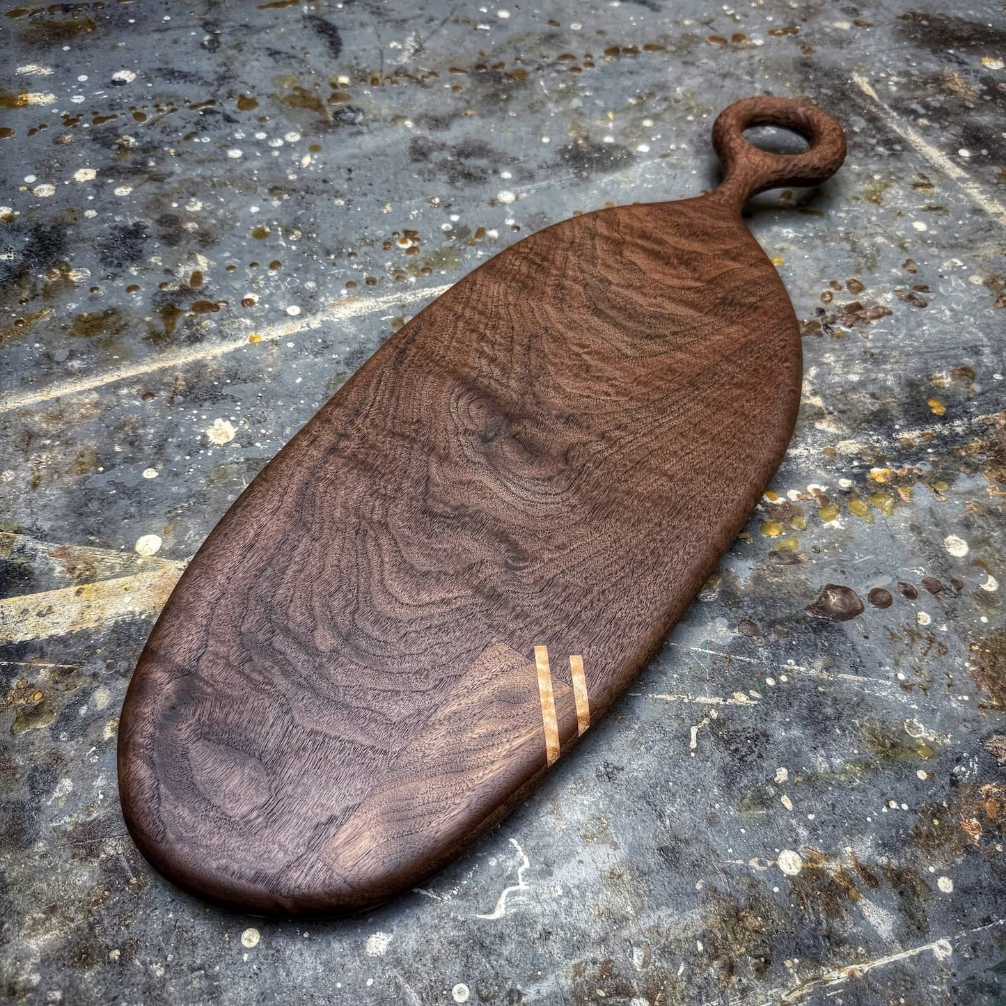 An oval wooden serving board with a loop handle, featuring rich dark brown wood grain and two small light-colored inlay stripes near the bottom, displayed on a textured, weathered gray stone surface.