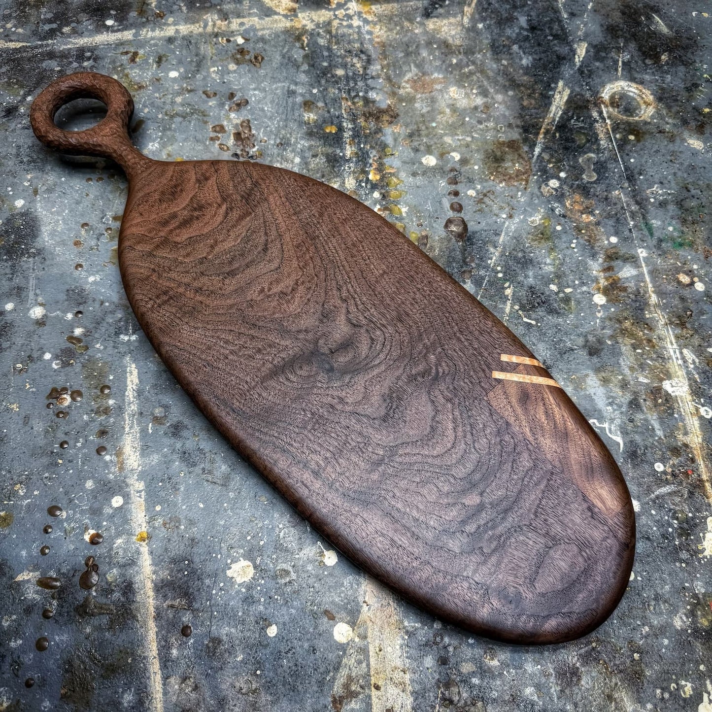 An organic-shaped wooden serving board with a loop handle, featuring a rich dark brown wood grain pattern and a small light-colored inlay accent, displayed on a textured, paint-splattered gray workbench.
