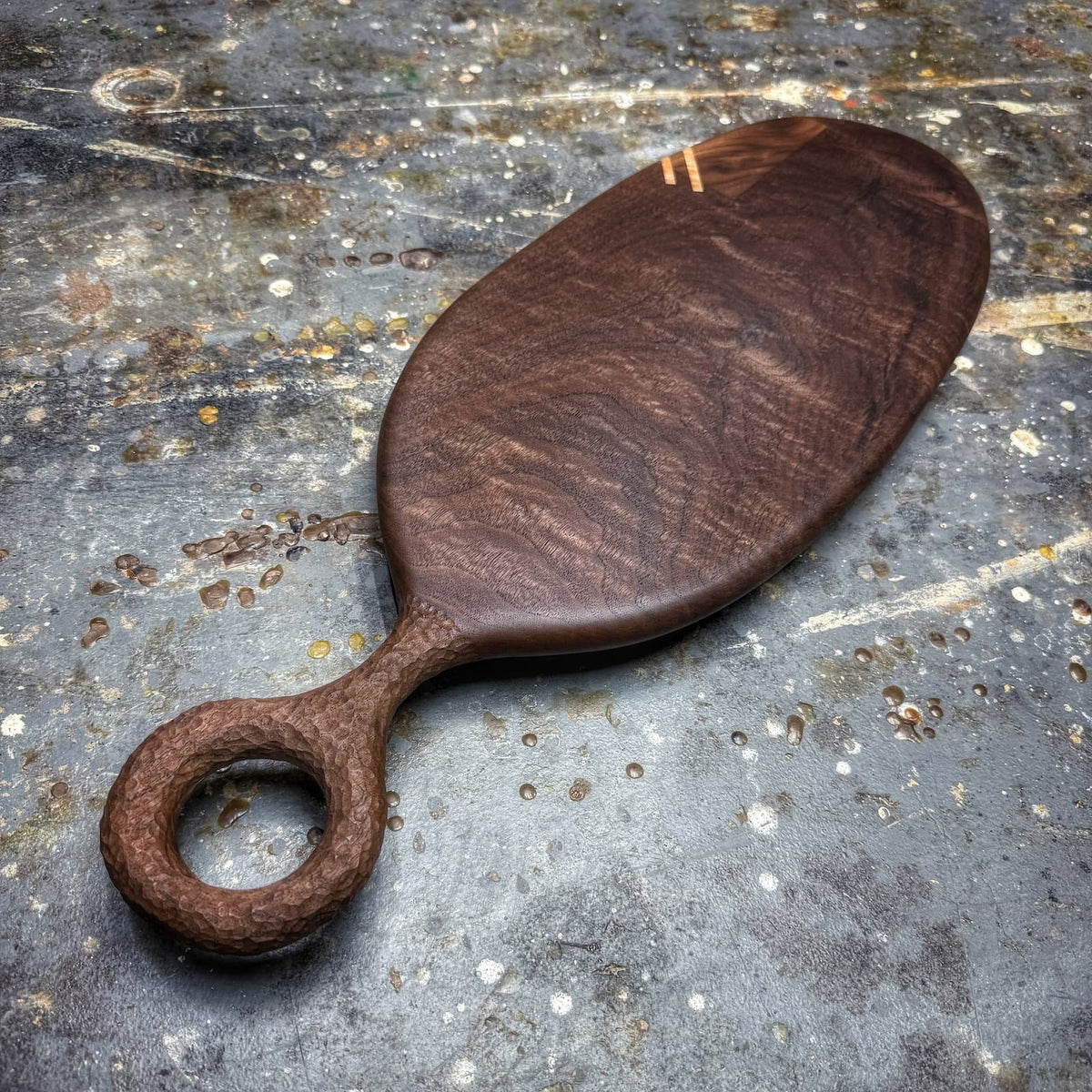 A dark brown wooden serving board with a loop handle, photographed on a textured, weathered gray stone surface.