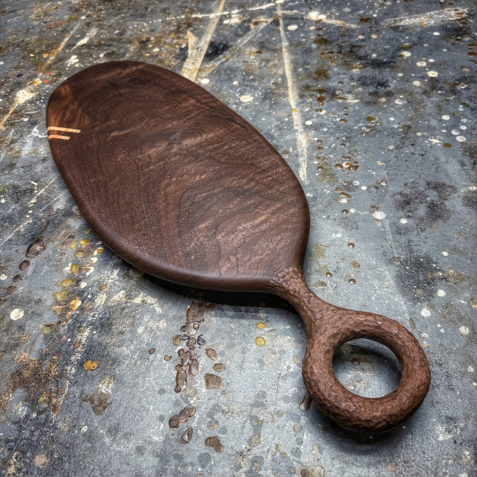 A dark brown wooden serving board with a loop handle, resting on a textured, weathered gray metal surface.