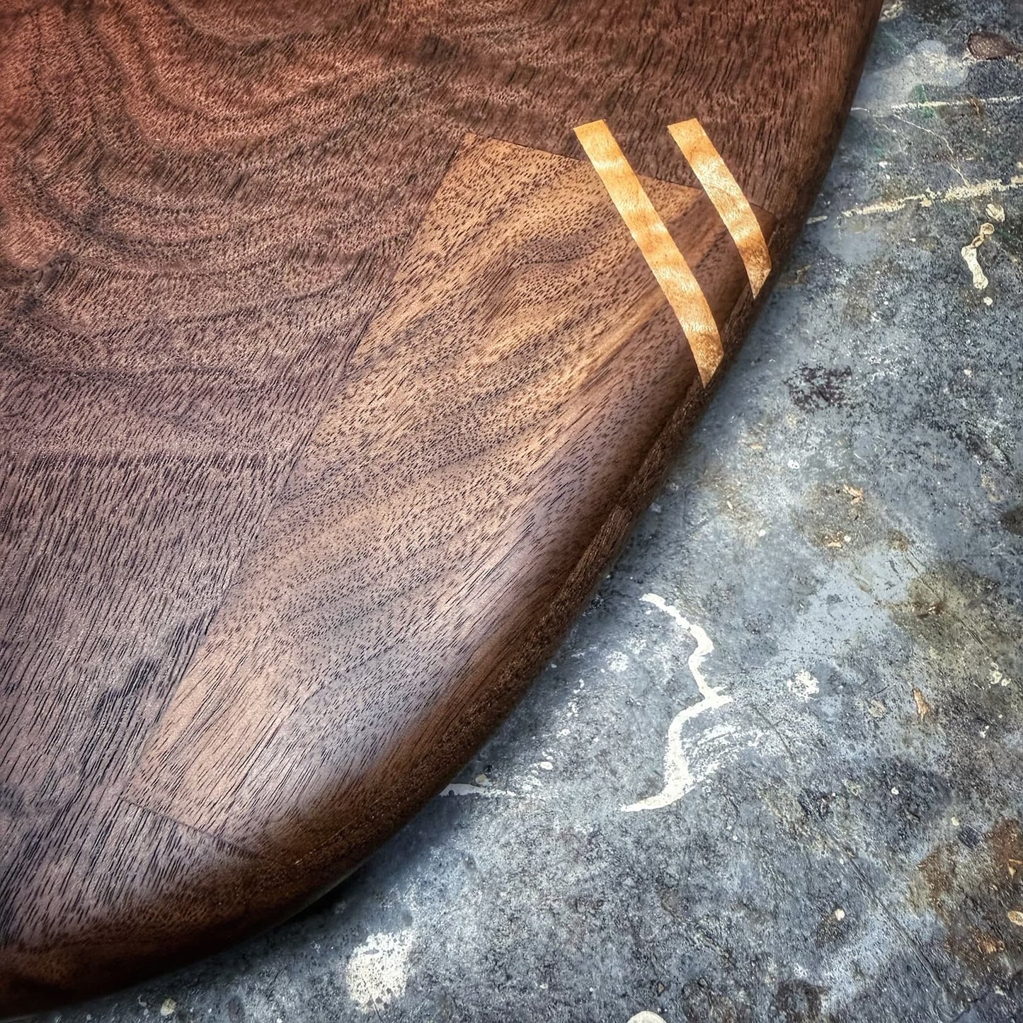 Close-up of a dark brown wooden board, likely a surfboard or paddle, showing a smooth wood grain finish and two light tan inlay stripes, resting on a textured gray concrete surface.