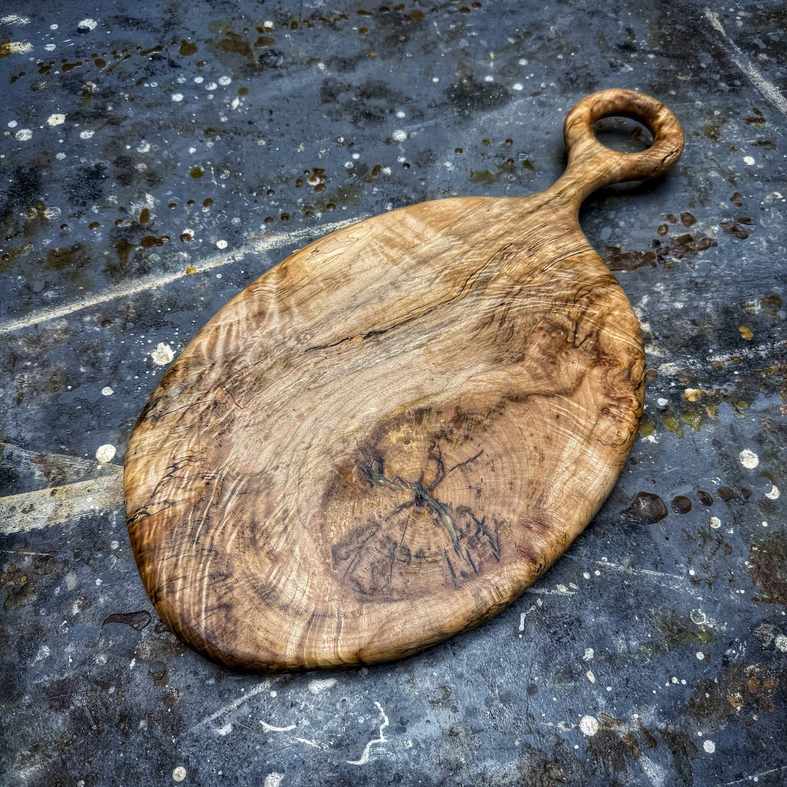 An organic-shaped wooden cutting board with a loop handle, resting on a dark, textured stone surface.