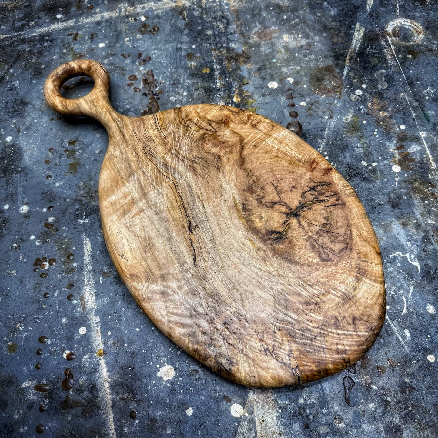 A rustic oval wooden cutting board with a loop handle, featuring natural wood grain and dark knots, displayed on a dark, weathered slate surface.