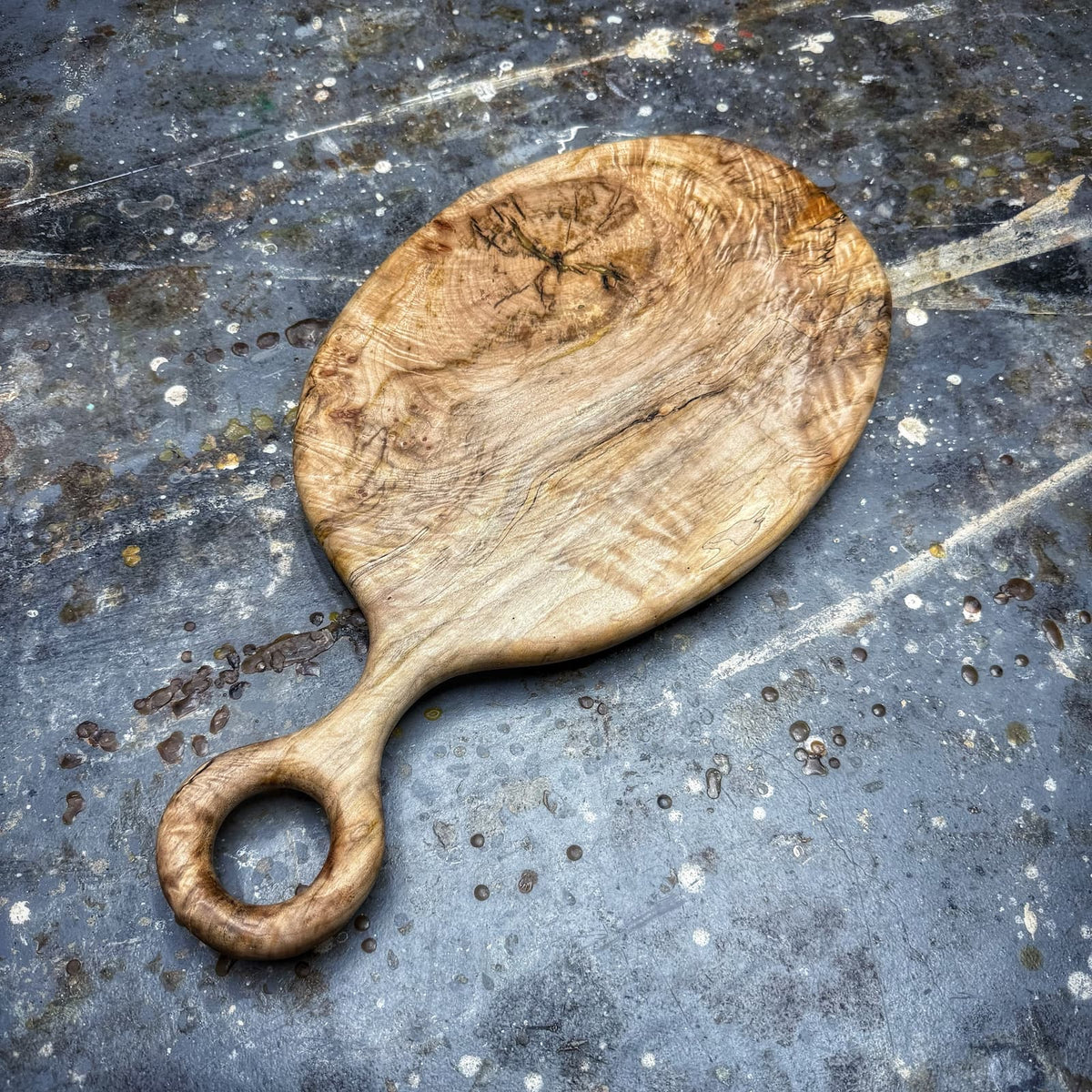 An oval wooden serving board with a loop handle, made from natural wood with visible grain and knots, placed on a dark textured slate surface.