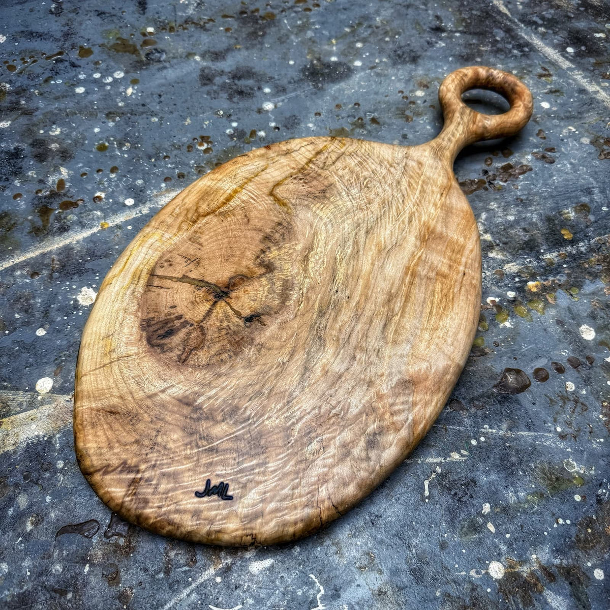 An oval wooden serving board with a loop handle, featuring natural wood grain and a small black painted mark near the bottom, placed on a dark textured stone surface.