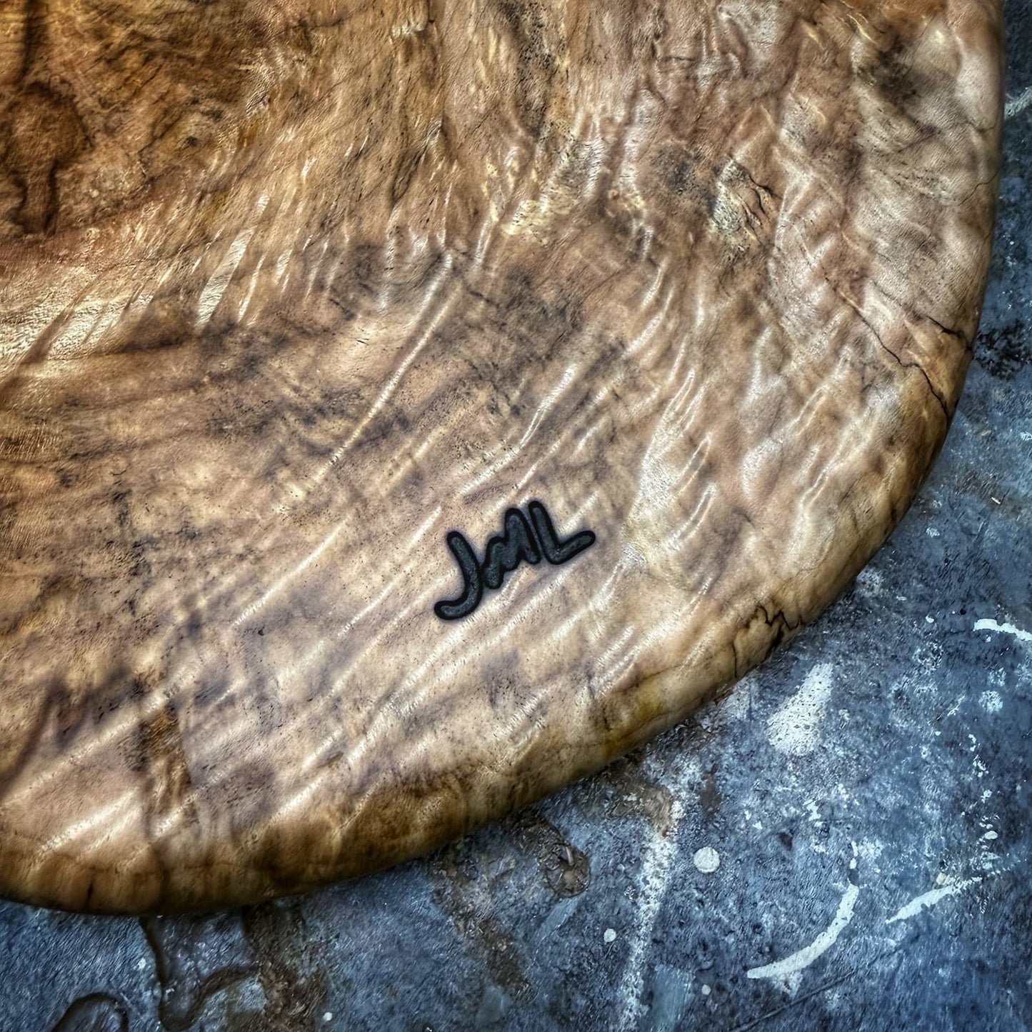 A close-up of a large, round wooden serving board with dramatic wavy grain patterns and a small black metal 'JML' logo, resting on a textured gray stone surface.